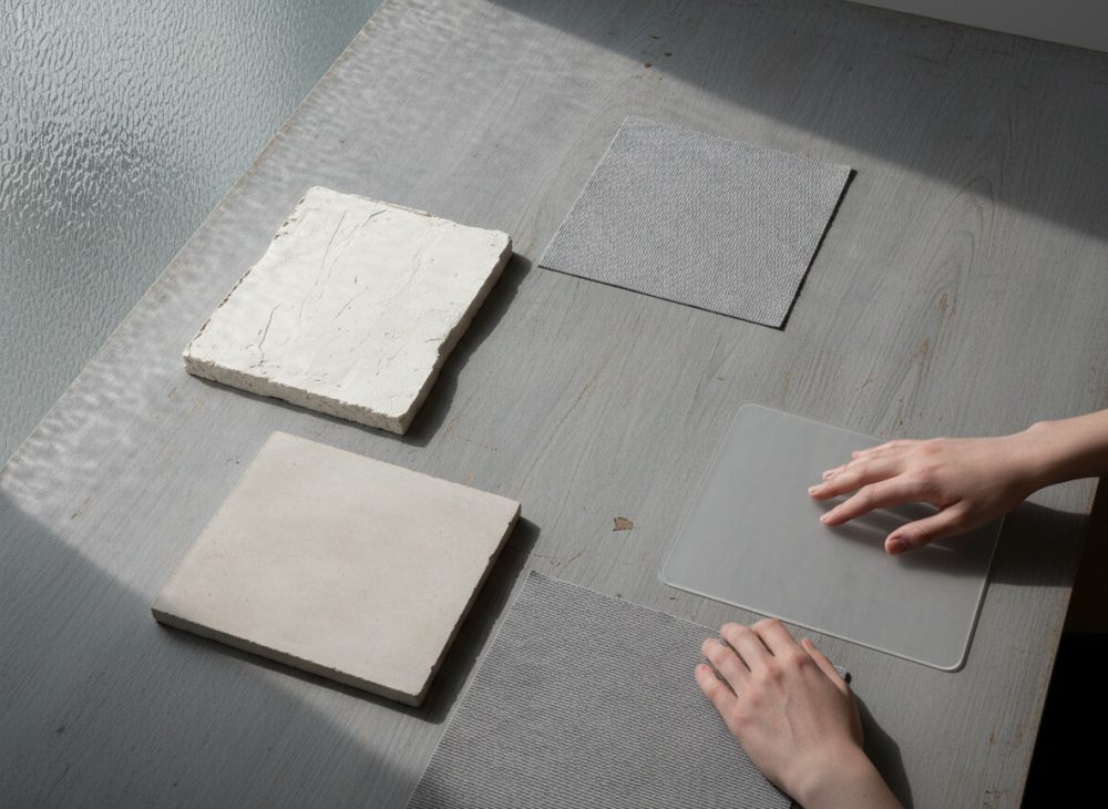 Amostras quadradas de superfícies neutras sobre mesa cinzenta com mãos a tocar amostra transparente