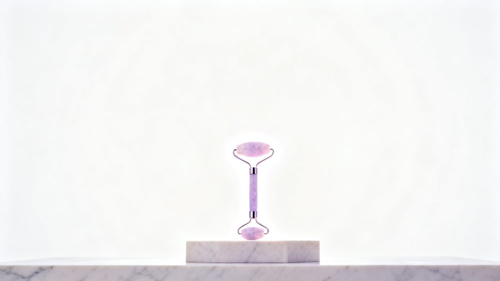 Rolo facial de quartzo rosa sobre suporte de mármore com fundo branco minimalista