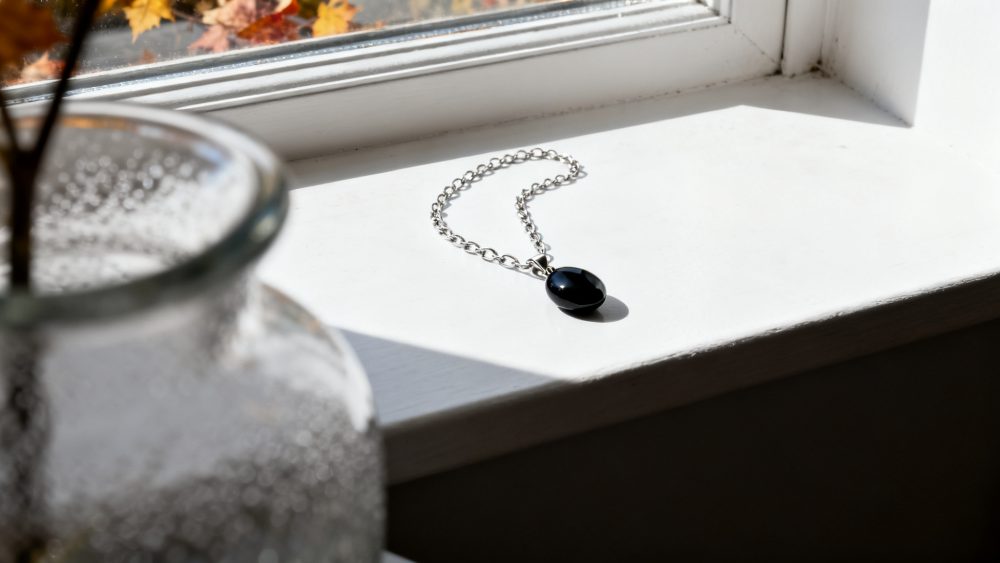 Colar prateado com pendente negro colocado num parapeito branco iluminado por luz natural