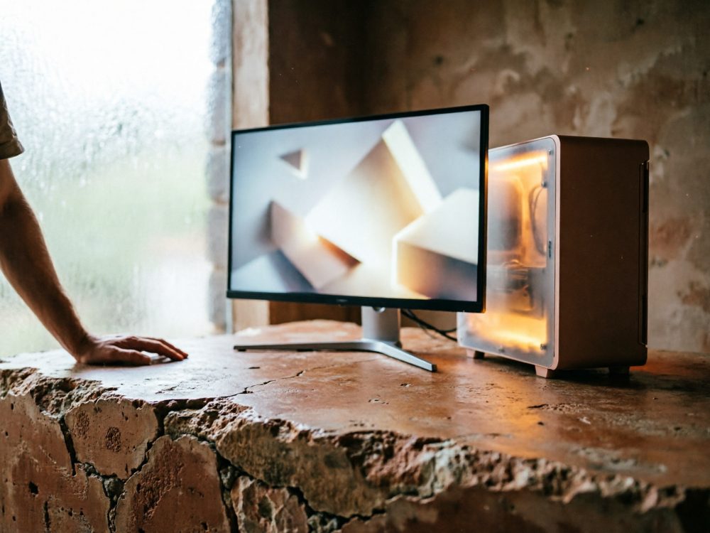 Computador gaming iluminado com monitor ultrafino numa mesa de pedra rústica perto de janela