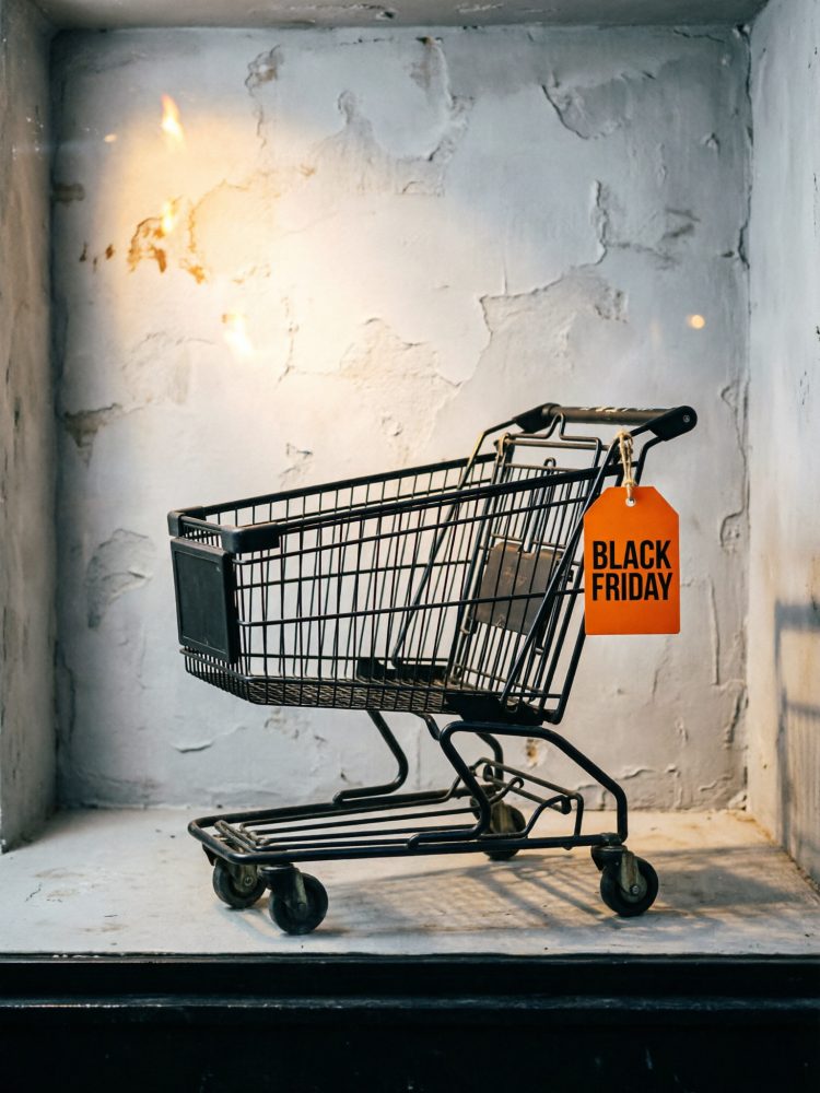 Carrinho de compras preto com etiqueta laranja Black Friday pendurada numa parede cinzenta texturada