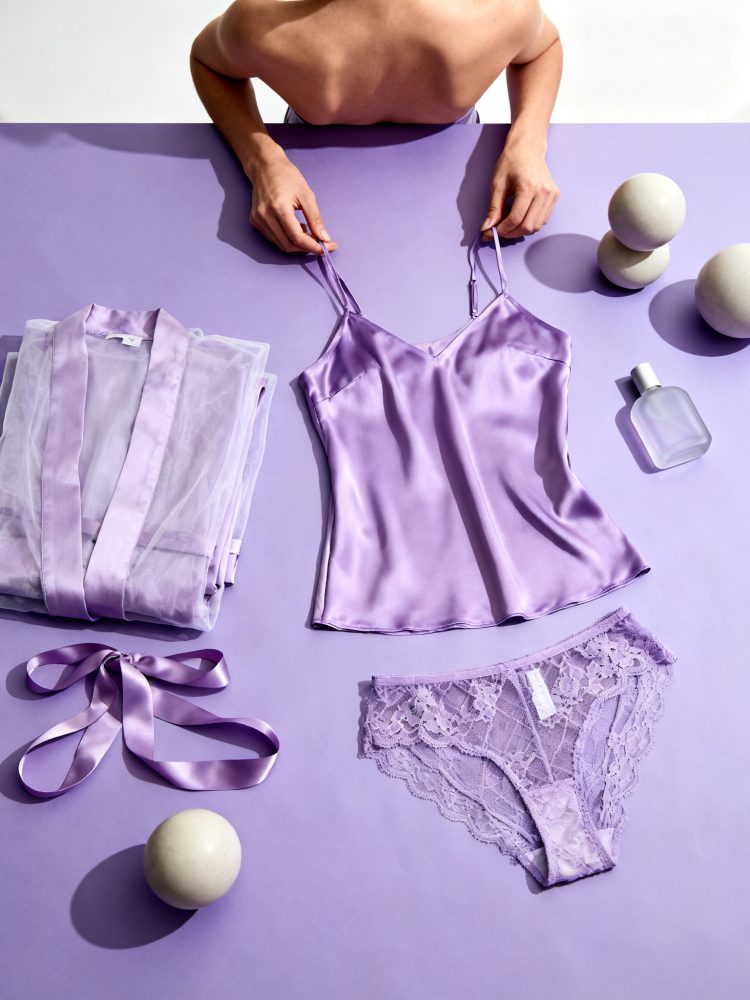 Conjunto de lingerie lilás com top de cetim, cueca de renda, robe transparente e fita sobre fundo lilás