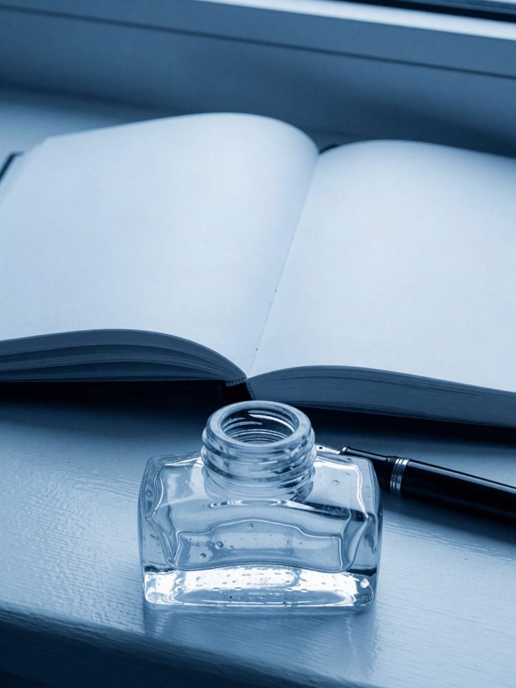 Caderno aberto com páginas em branco, tinteiro de vidro e caneta preta sobre uma superfície azulada