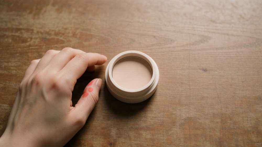 Mão feminina junto a pote branco com base de maquilhagem cremosa sobre mesa de madeira