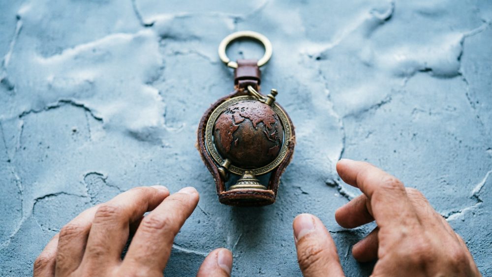 Chaveiro de mini globo terrestre metálico e couro sobre mesa cinza com mãos próximas