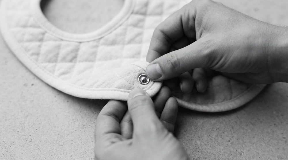 Babete acolchoado branco com fecho de pressão a ser apertado por duas mãos.