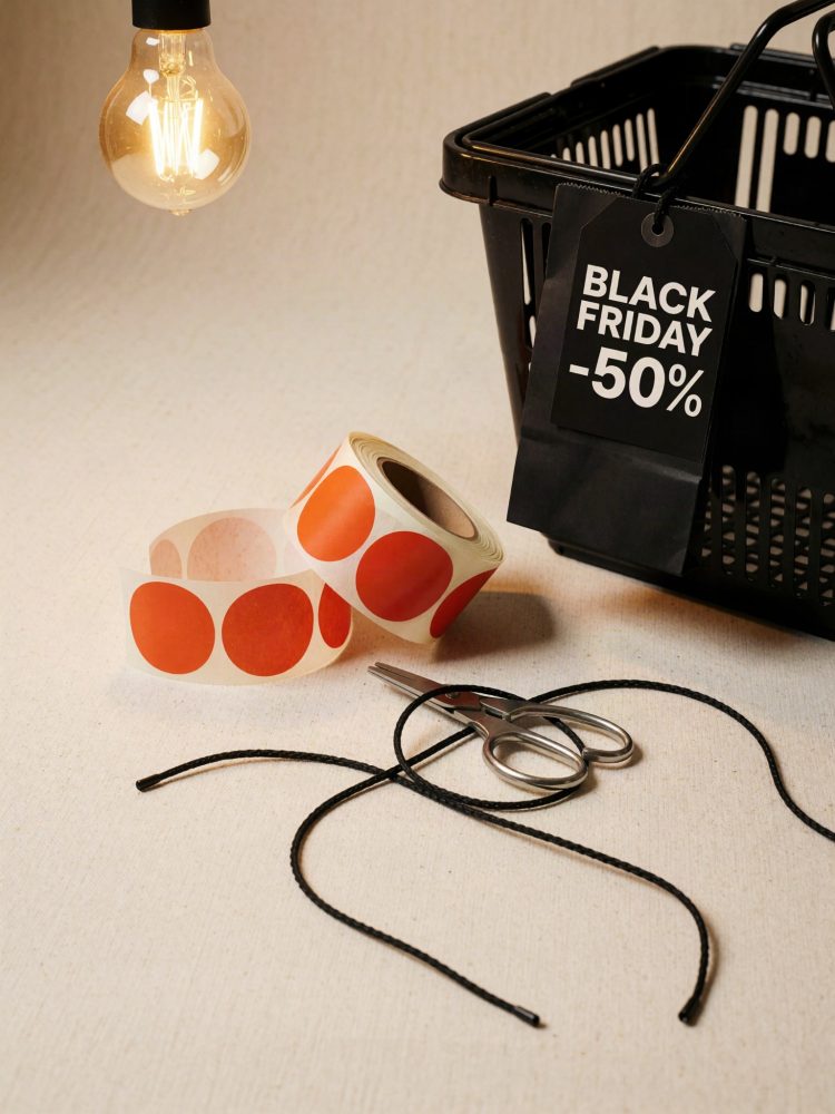 Cesto preto com etiqueta Black Friday -50%, rolos de etiquetas vermelhas, tesoura e fio preto sobre mesa clara