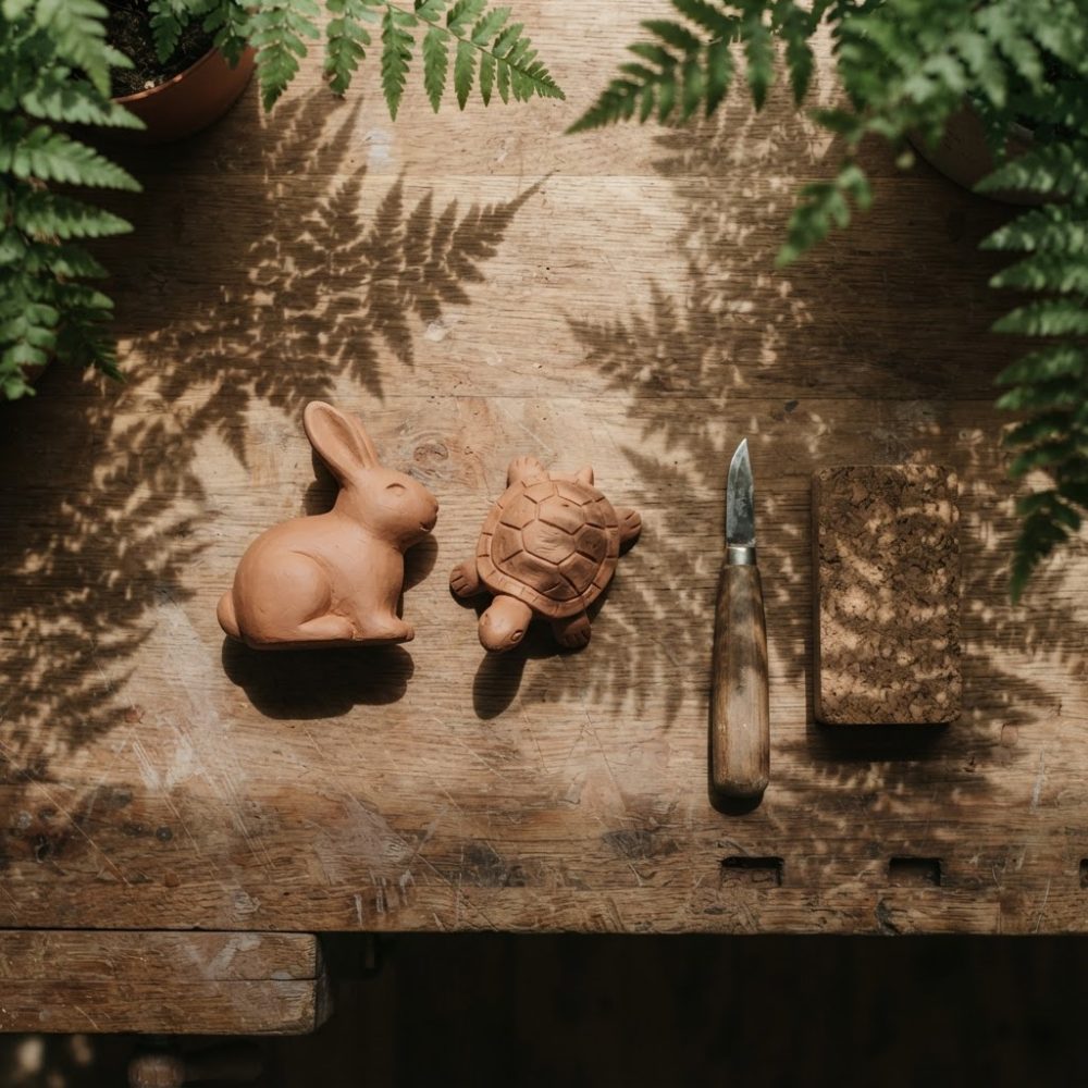 Brinquedos de cerâmica em forma de coelho e tartaruga sobre mesa de madeira com sombras de folhas