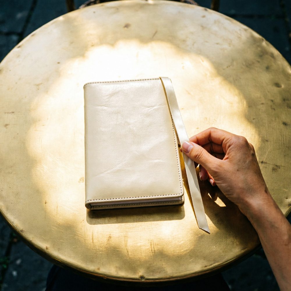 Caderno bege em pele com marcador de fita sobre mesa dourada redonda, com mão segurando o marcador