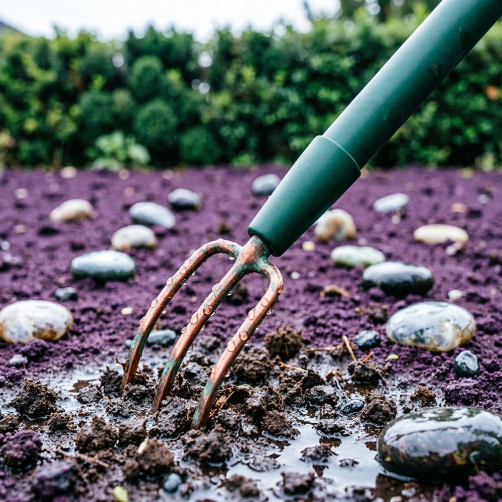 Garfo de jardinagem a revolver solo húmido escuro com pedras e vegetação desfocada ao fundo