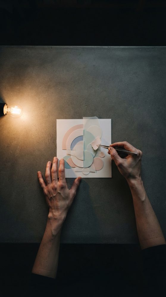 Mãos a trabalhar em arte abstrata com formas circulares pastel sobre mesa cinza iluminada por luz quente