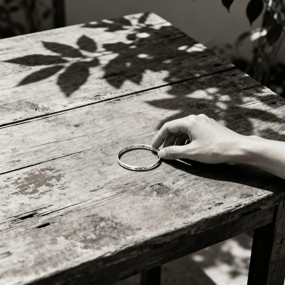 Mão segurando pulseira de prata sobre mesa de madeira com sombras de folhas