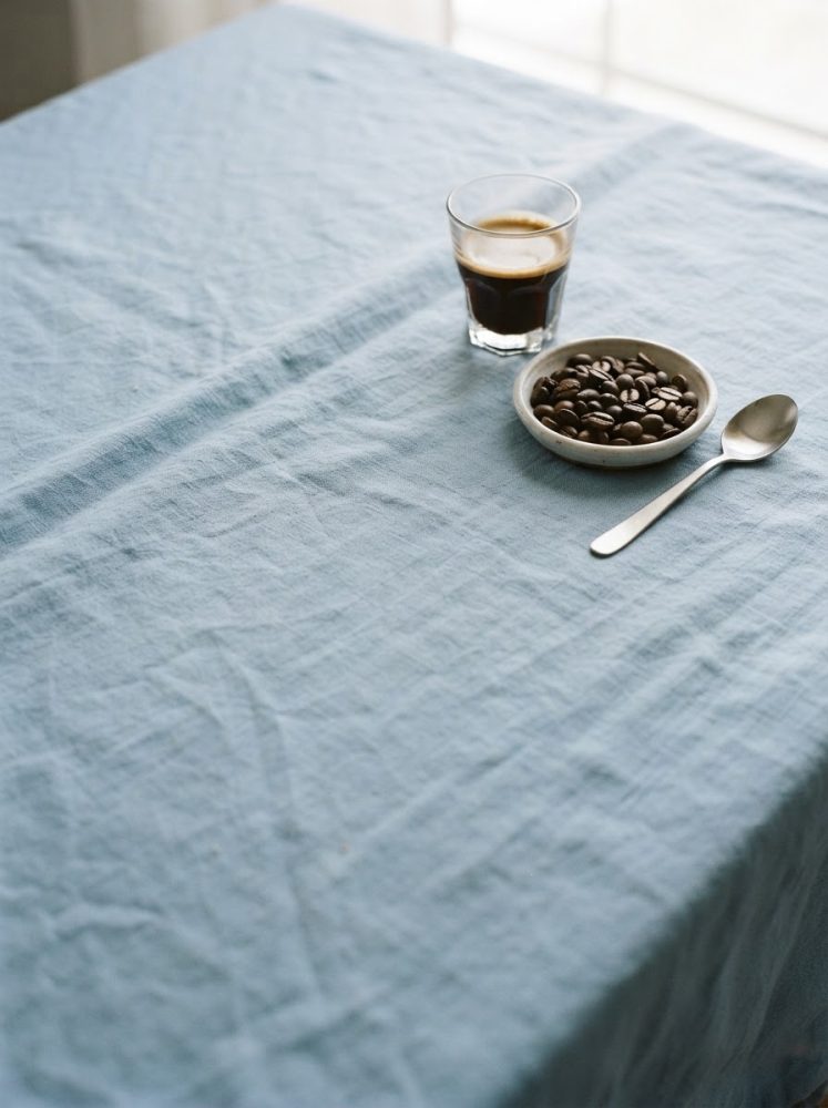 Copo de café espresso, taça com grãos de café e colher numa mesa azul clara com luz natural