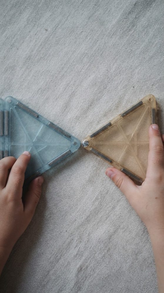 Duas mãos infantis a segurar peças triangulares magnéticas azul e bege sobre tecido cinzento