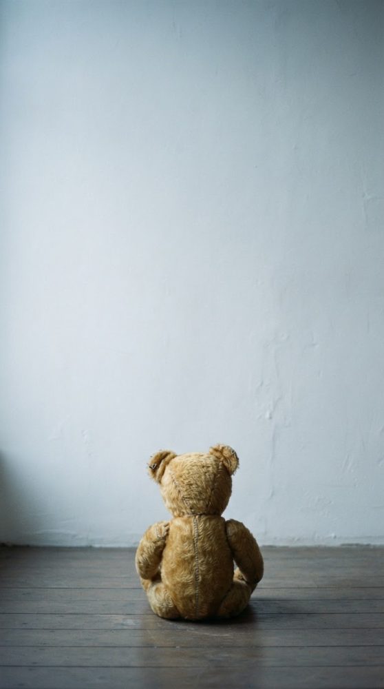 Ursinho de peluche castanho vintage sentado de costas sobre chão de madeira com parede branca ao fundo
