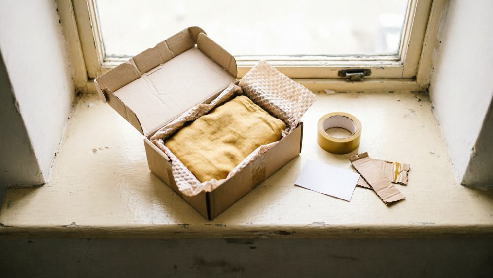 Caixa de cartão aberta com tecido amarelo protegido por papel bolha, fita adesiva e etiqueta em cima de uma superfície clara junto à janela