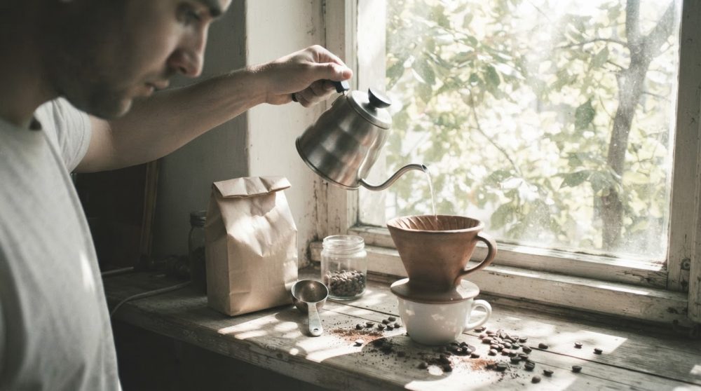 Homem a preparar café coado com bule metálico junto a janela com luz natural e grãos espalhados