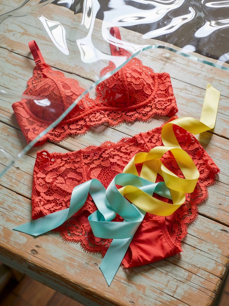 Conjunto vermelho de lingerie em renda com fitas amarelas e azuis sobre mesa de madeira