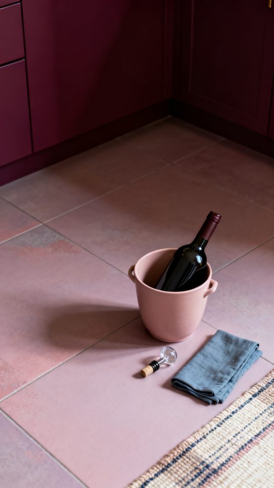 Garrafa de vinho tinto num balde de cerâmica rosa com guardanapo azul ao lado no chão de cozinha