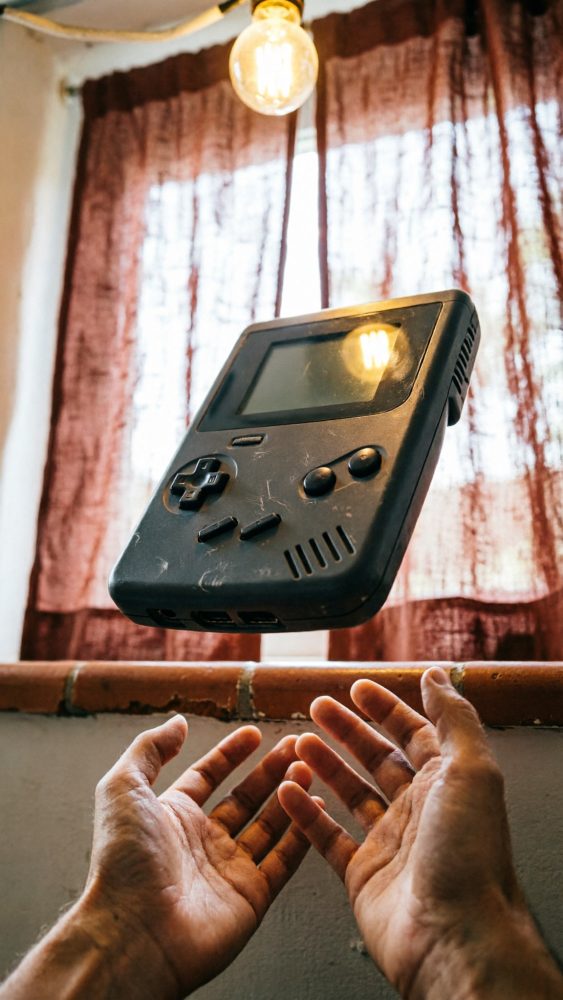 Consola de jogos portátil preta flutuando sobre mãos abertas junto a janela com cortinas castanhas