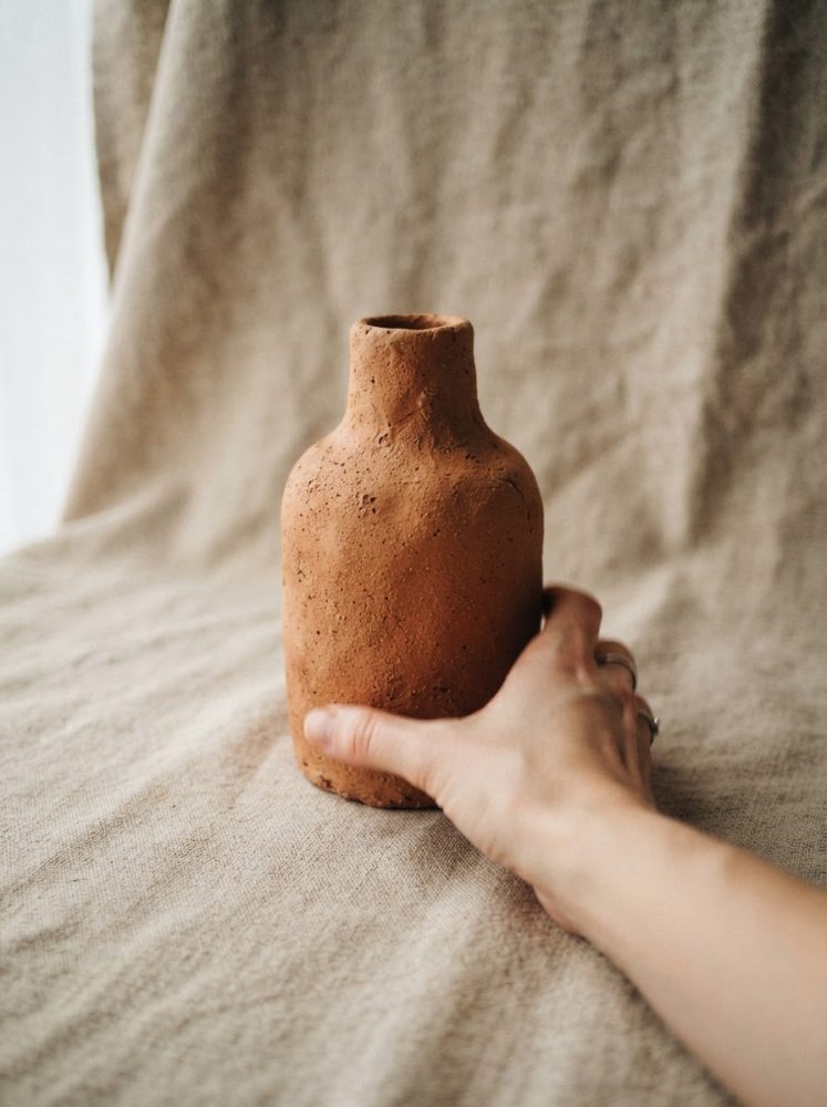 Vaso em terracota colocado sobre tecido bege texturizado com mão segurando base