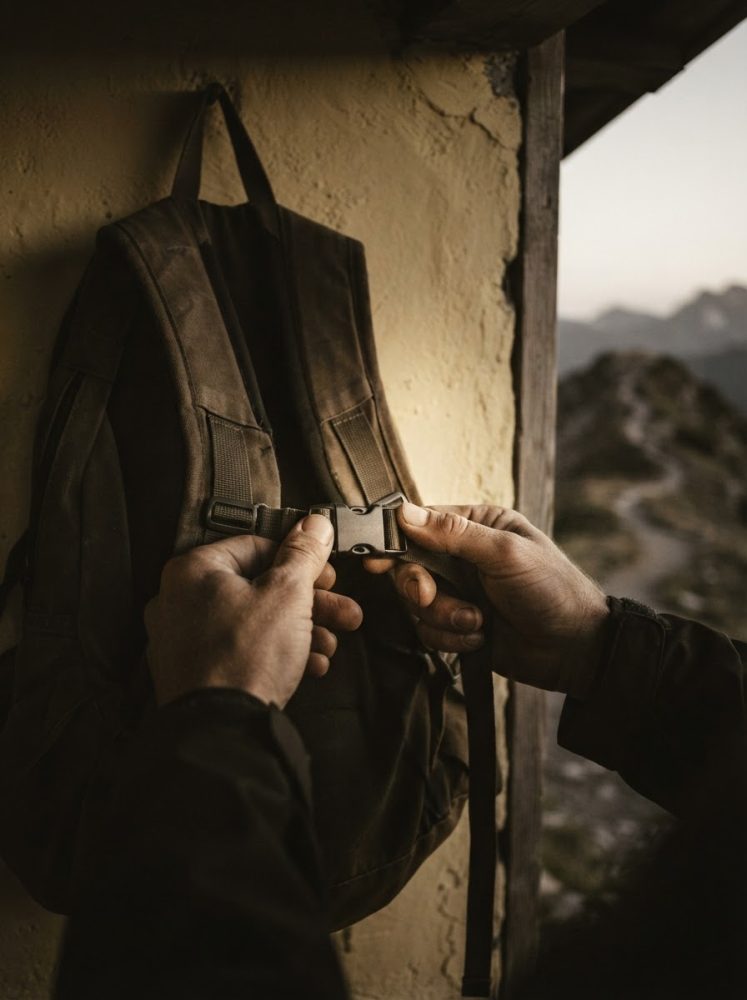 Mochila castanha pendurada numa parede com mãos a fechar fecho, vista montanha ao fundo