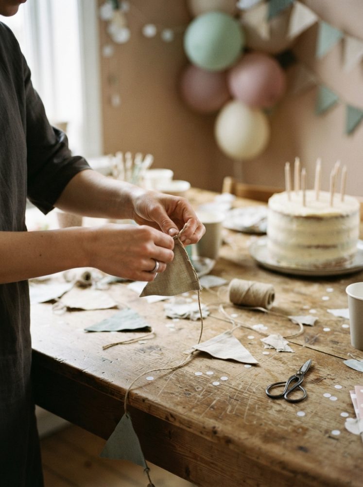Mãos a montar bandeirolas de tecido sobre mesa de madeira com bolo e balões ao fundo
