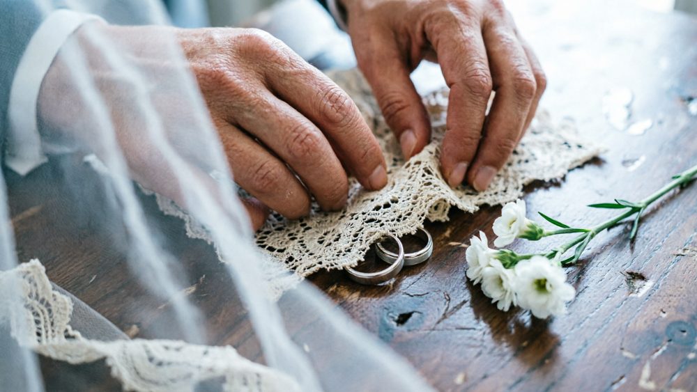 Mãos ajustando renda delicada sobre alianças de casamento e flores brancas numa mesa de madeira