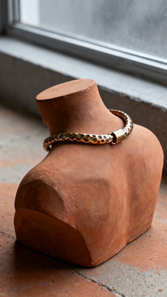 Colar dourado martelado num busto de terracota junto a uma janela com luz natural