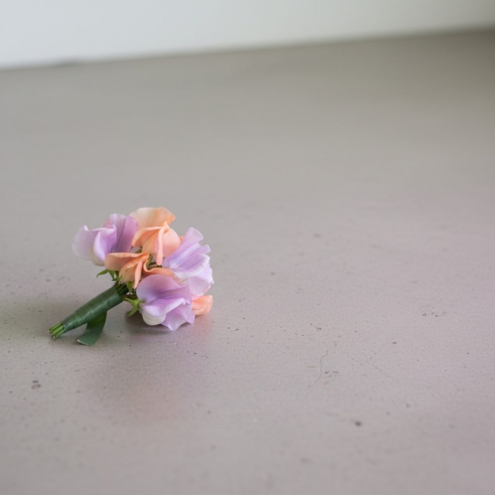 Mini buquê com flores lilás e pêssego atado com fita verde sobre superfície cinza