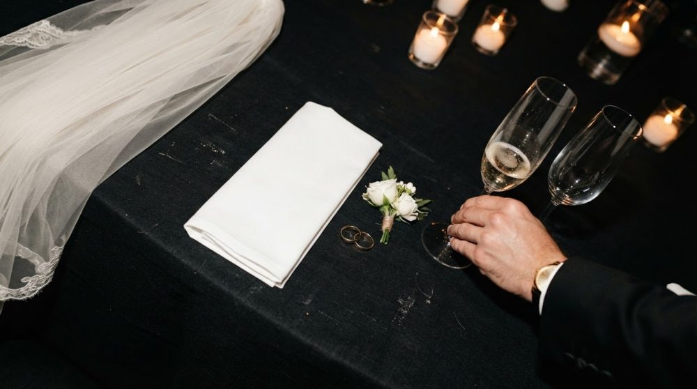 Mesa com véu de noiva, guardanapo, alianças, boutonnière, taças de champanhe e velas acesas
