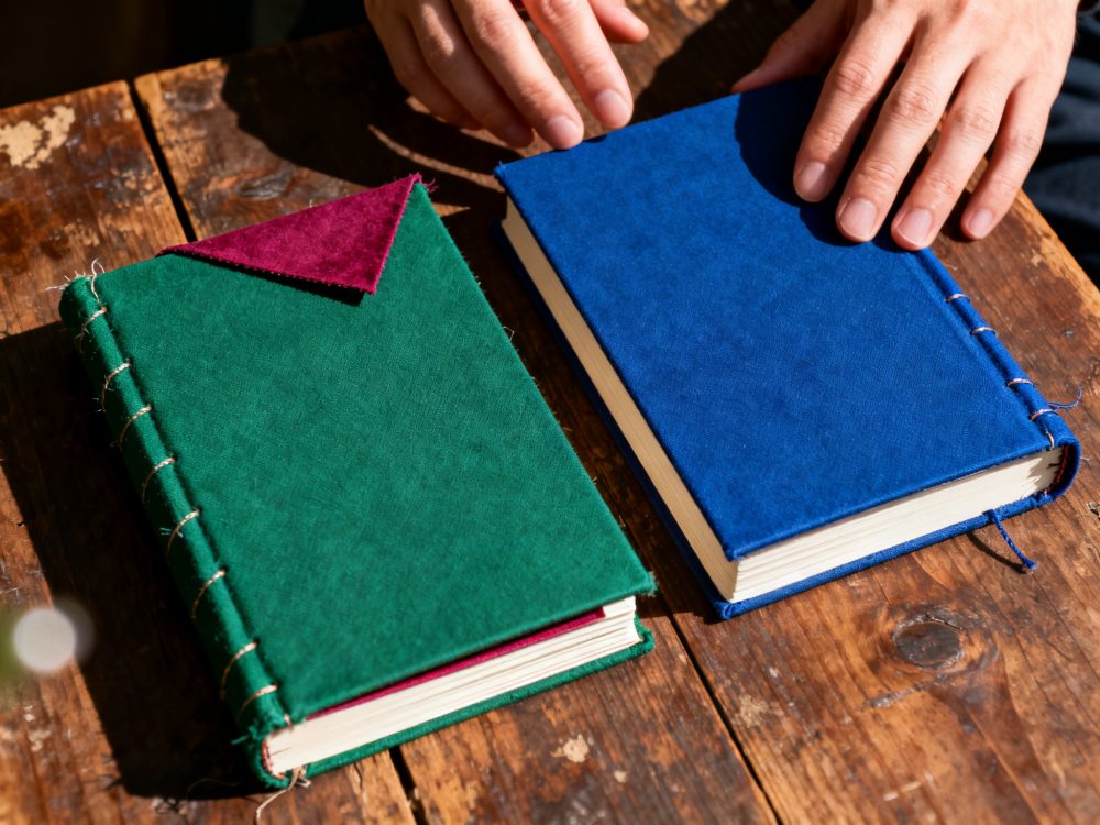 Dois livros artesanais em tecido azul e verde sobre mesa de madeira com mãos a tocar o livro azul