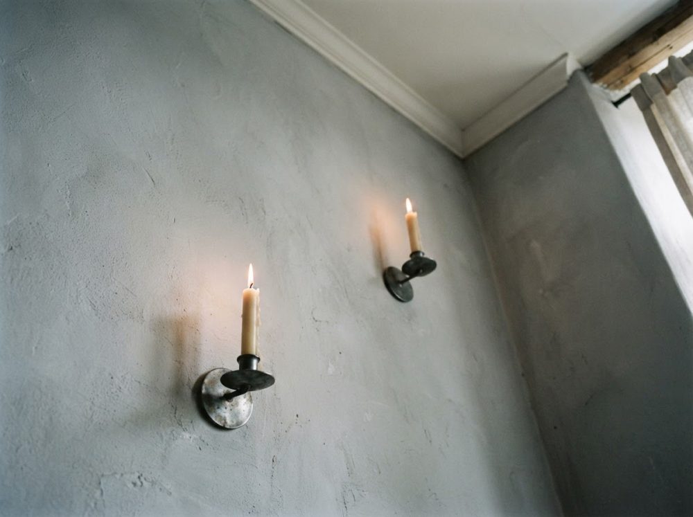 Duas velas finas acesas em suportes metálicos numa parede cinza com textura