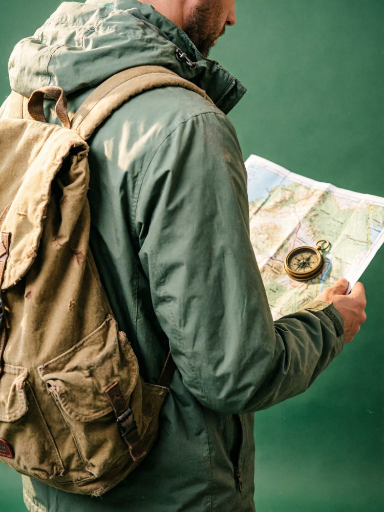 Pessoa de costas com casaco verde e mochila castanha segura mapa com bússola dourada em fundo verde