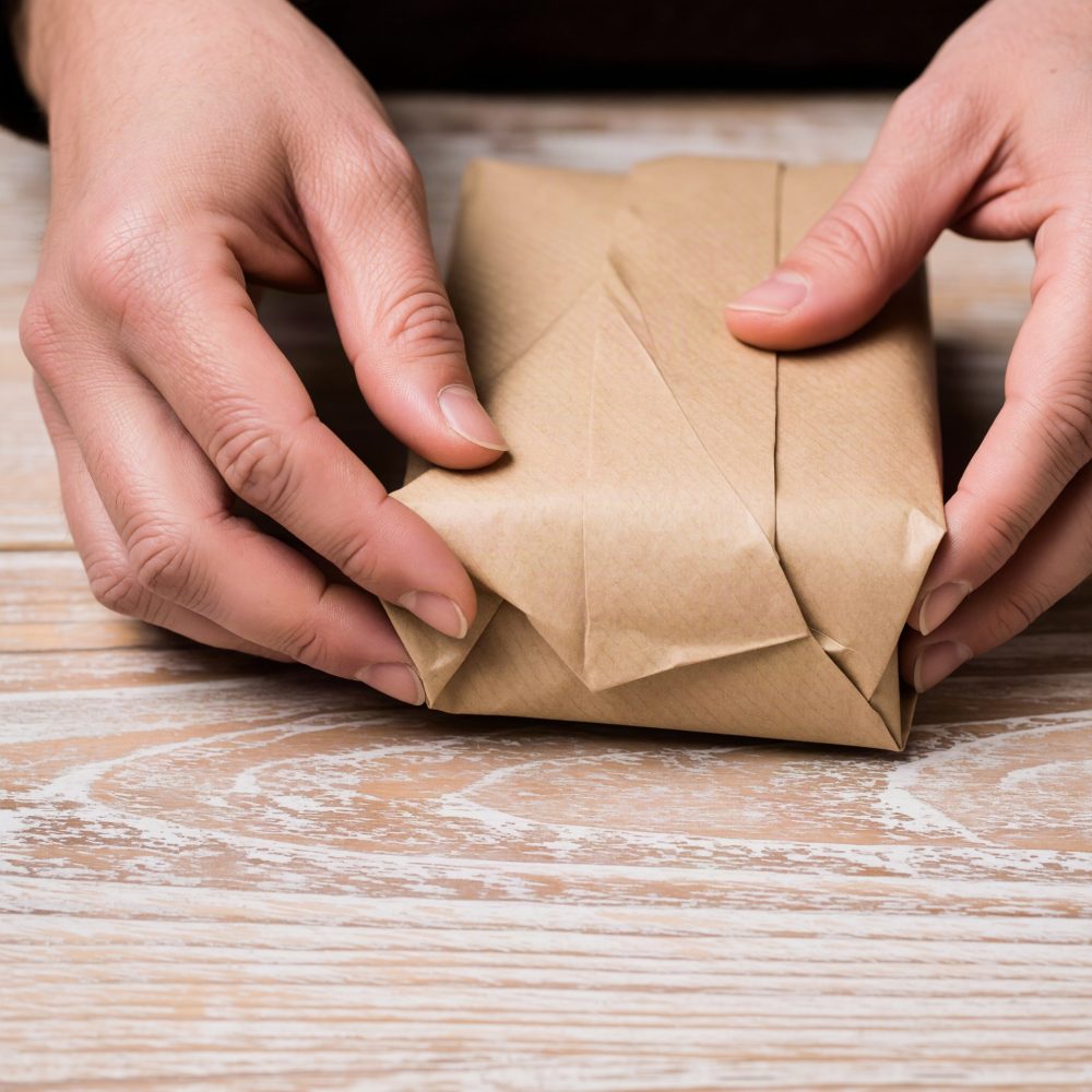 Mãos a embrulhar um pacote pequeno com papel kraft castanho numa mesa de madeira clara