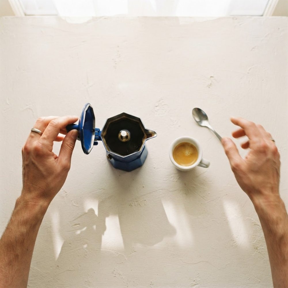 Mãos a abrir tampa de cafeteira moka azul com café expresso e colher numa mesa branca