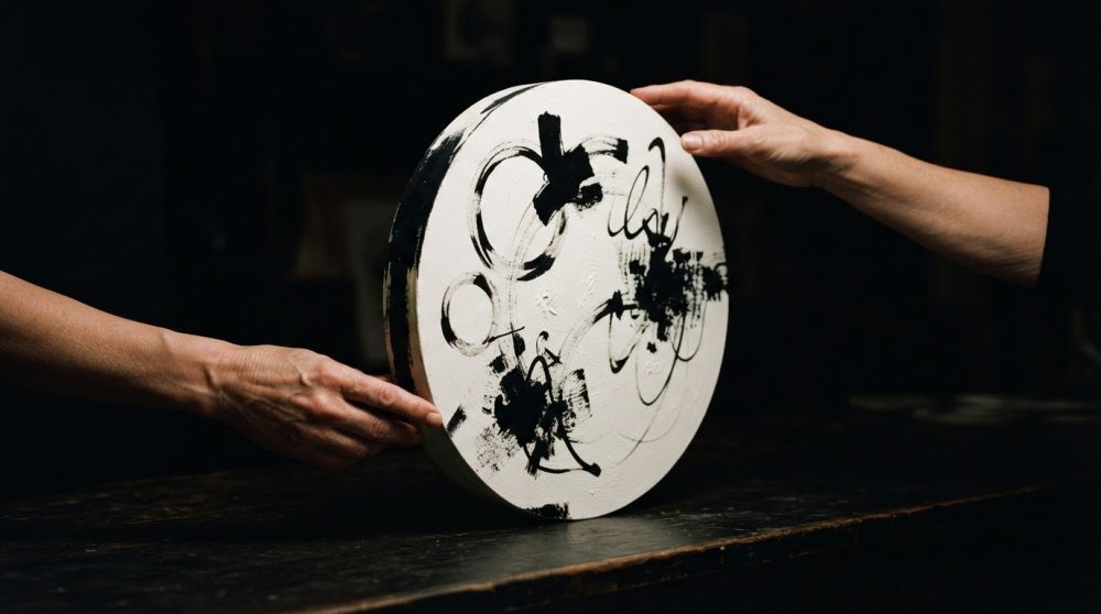 Duas mãos seguram uma tela circular com pintura abstrata preta sobre fundo branco em ambiente escuro