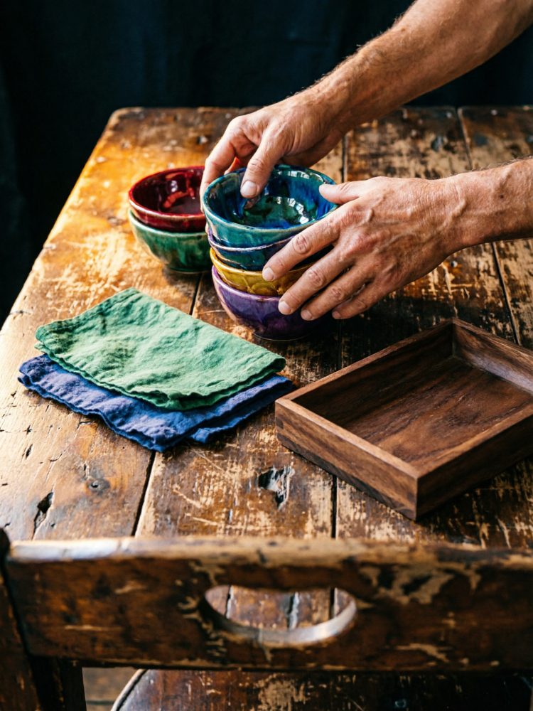 Mãos empilhando taças coloridas sobre mesa de madeira rústica com guardanapos e caixa de madeira