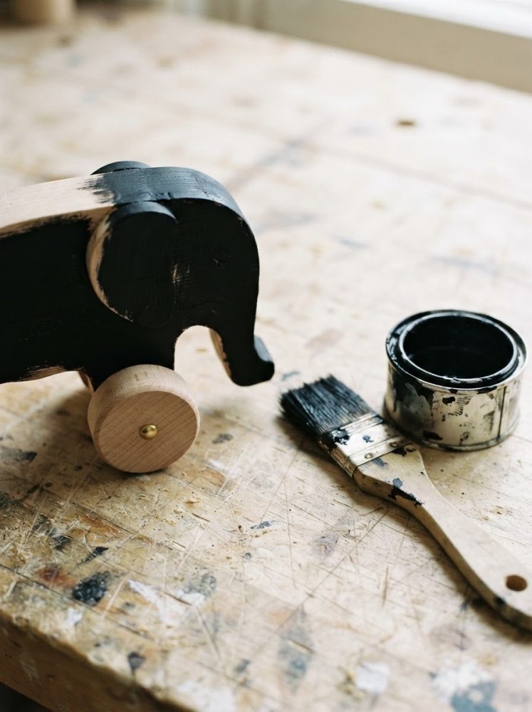 Brinquedo elefante de madeira pintado de preto com pincel e tinta preta sobre banco de trabalho rústico.