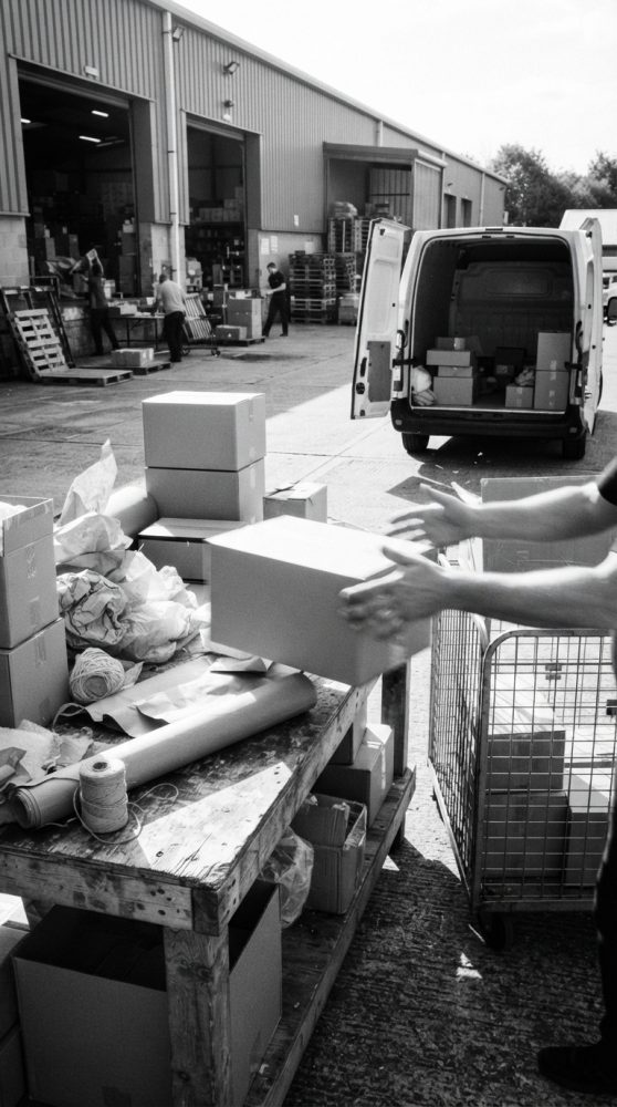 Pessoa a embalar caixas de cartão numa mesa fora de armazém com carrinha de transporte ao fundo