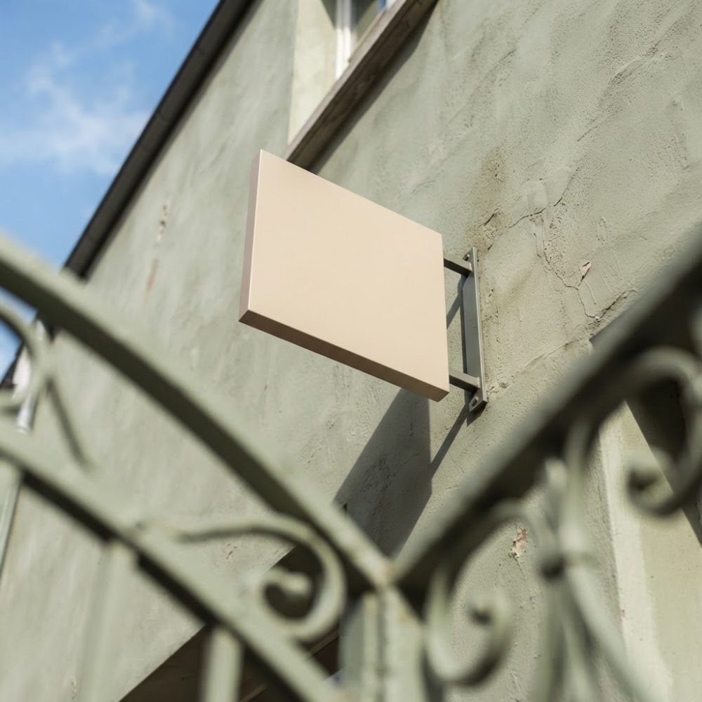 Placa retangular bege em suporte metálico na parede exterior de loja com corrimão verde desfocado