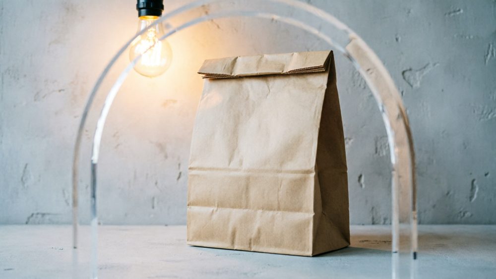 Saco de papel kraft fechado sobre mesa com lâmpada pendurada e fundo cinza texturizado