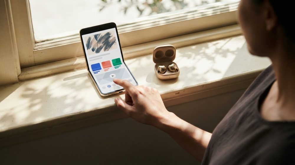 Pessoa manuseia smartphone dobrável junto de auscultadores sem fios dourados numa janela iluminada