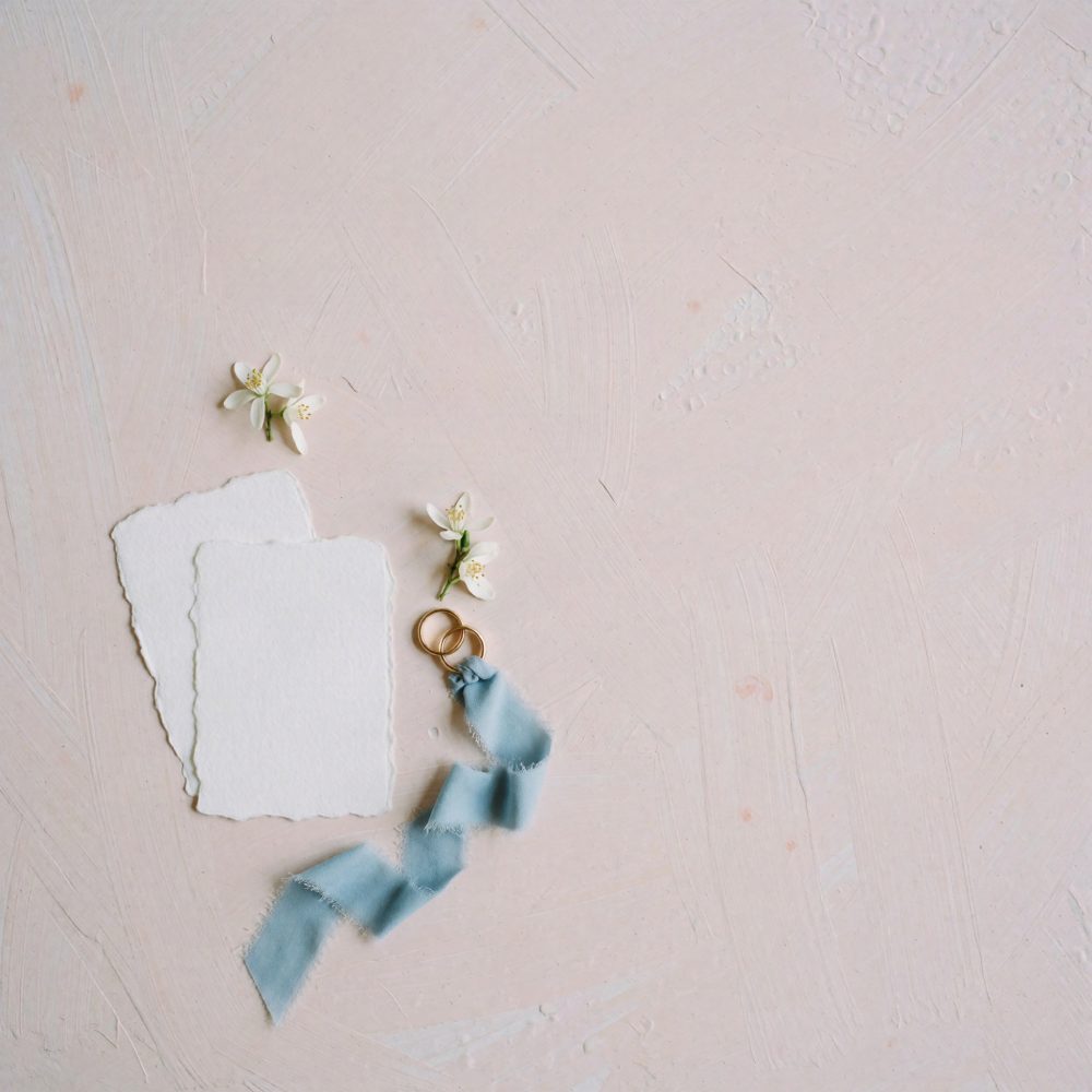 Convite de casamento branco com flores, alianças douradas e fita azul sobre fundo rosa texturizado