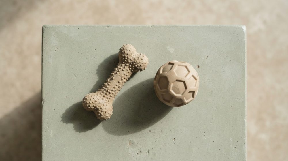 Dois brinquedos para cães, um osso e uma bola em tons neutros sobre superfície cinza