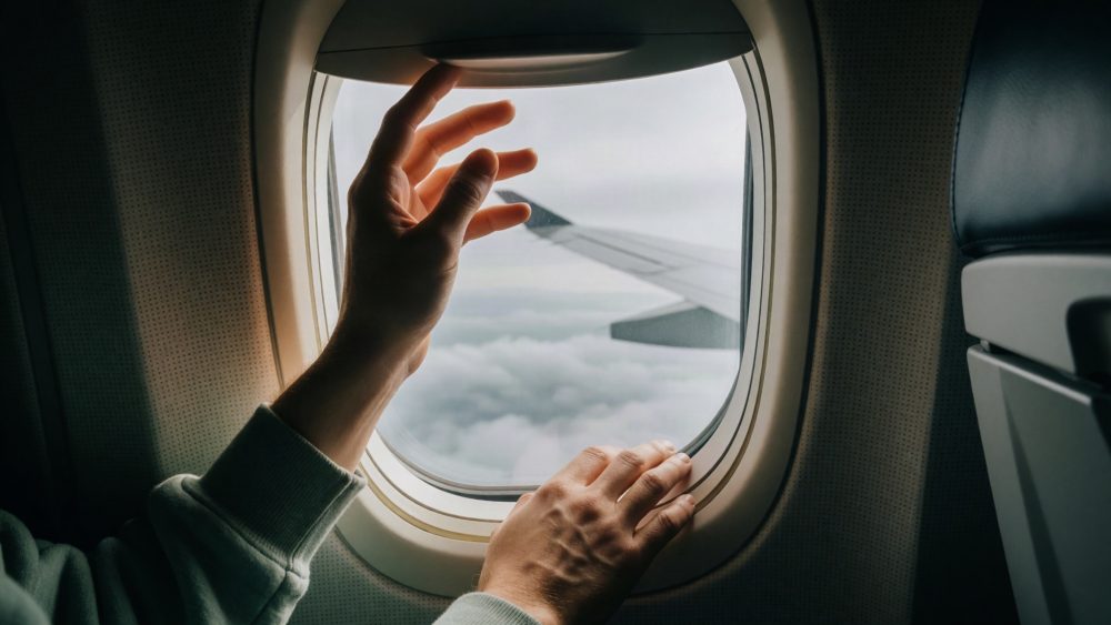 Mãos a abrir persiana de janela de avião com vista para asa e nuvens no exterior