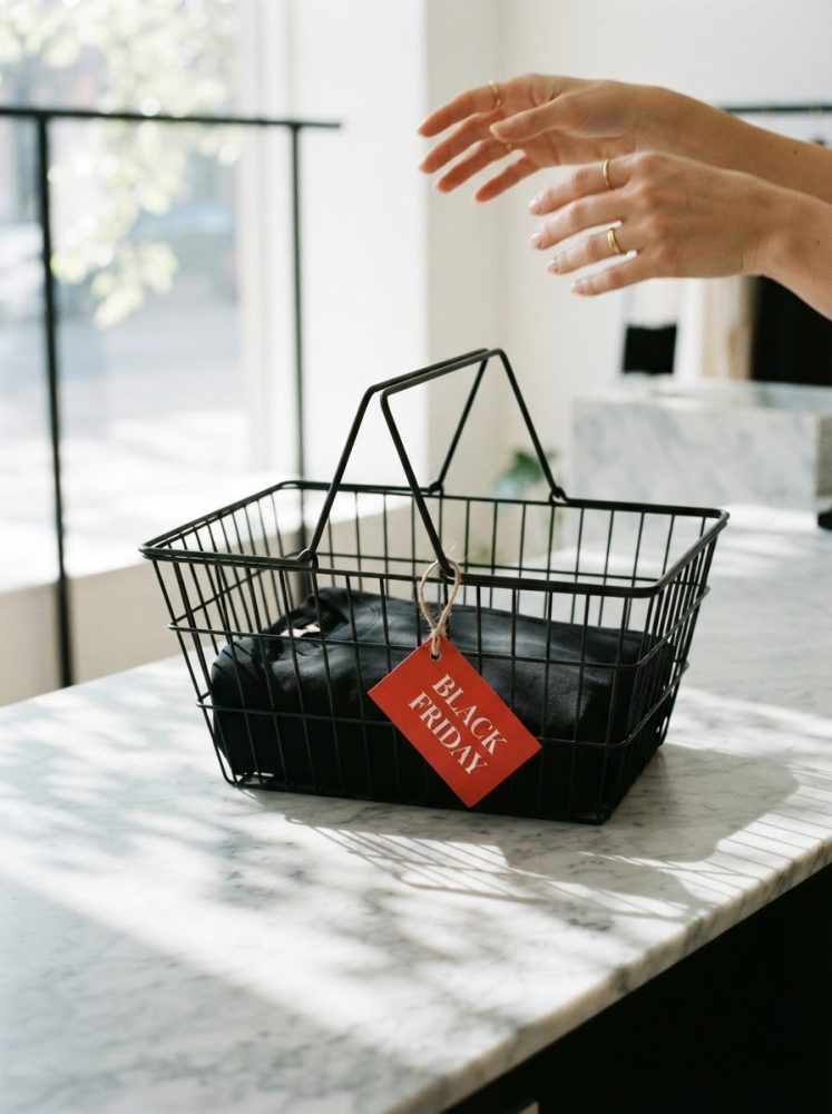 Mãos junto a um cesto de compras preto com etiqueta vermelha Black Friday numa bancada de mármore branco