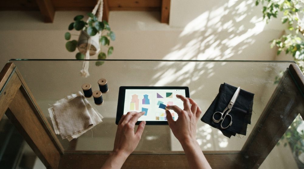 Mãos usam tablet para design de moda digital sobre mesa com tecidos, tesoura e linhas