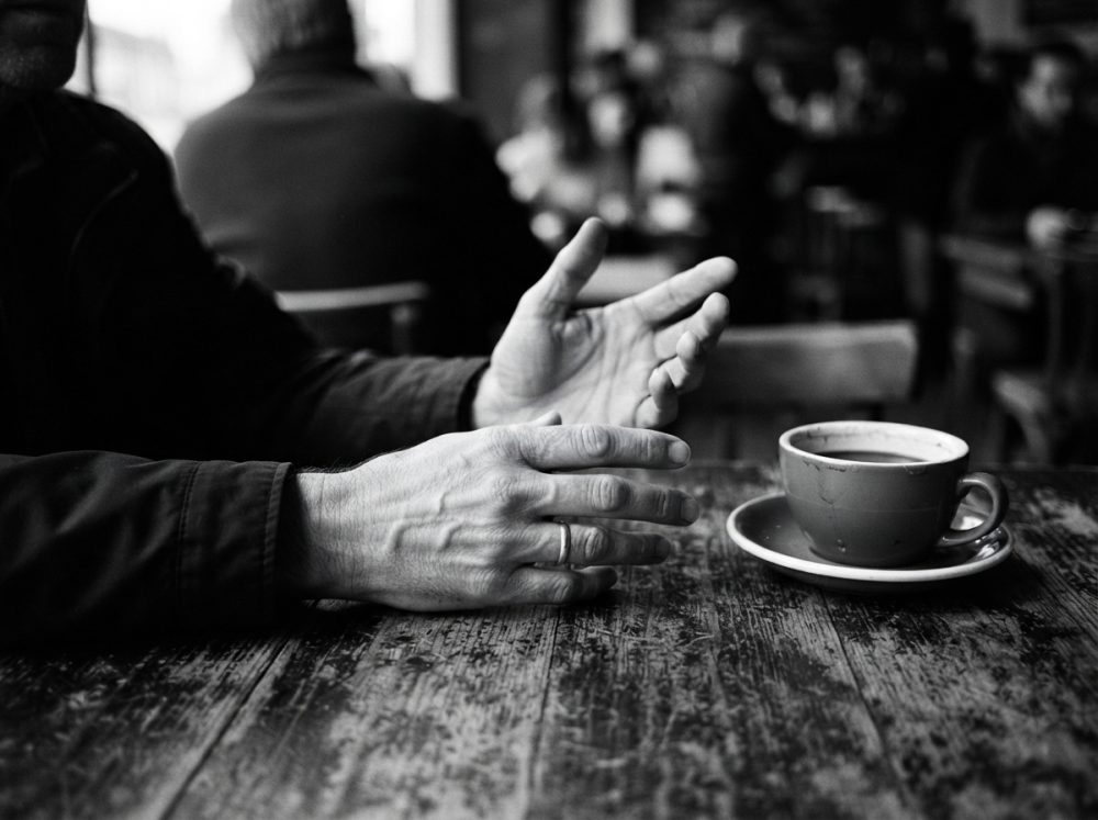 Mãos expressivas junto a chávena de café sobre mesa rústica num ambiente de café