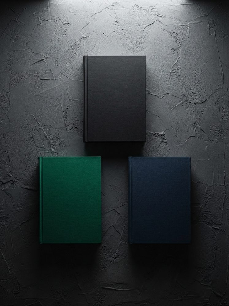 Três livros de capa dura preta, verde e azul sobre fundo cinza texturado